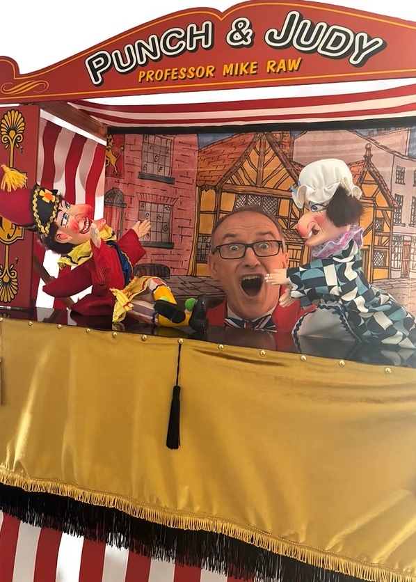 Michael Raw performing as Mr PunchandJudy, standing beside a traditional red‑and‑white Punch & Judy booth, smiling while entertaining children with puppets and balloon model